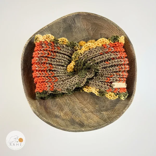 Hand-knitted wide acrylic headband laid flat on a wooden plate, forest green, orange,  and dark yellow gradient with twisted front design, ribbed knit texture and hand-crocheted scalloped edge, RAHEcraft