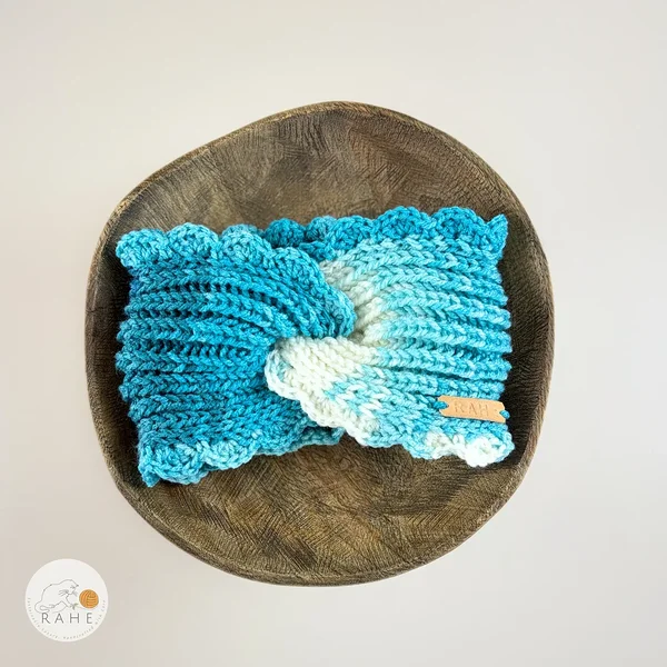 Hand-knitted wide acrylic headband laid flat on a wooden plate, white  and turquoise blue gradient with twisted front design, ribbed knit texture and hand-crocheted scalloped edge, RAHEcraft