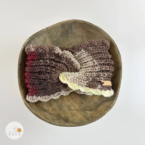 Hand-knitted wide acrylic headband laid flat on a wooden bowl, twisted design, brown, beige and red tones, crocheted scalloped lace edge, RAHEcraft