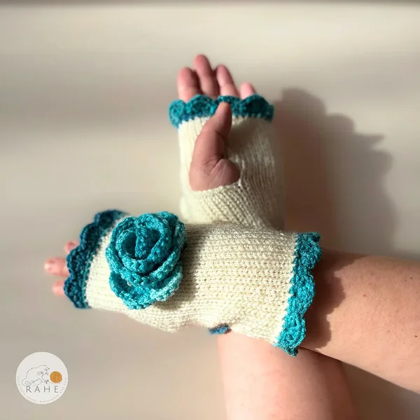 Close-up of RAHEcraft hand-knitted fingerless gloves in ivory white. The textured crochet flower and deep teal lace edging create a romantic and feminine look.