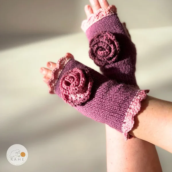 Close-up of RAHEcraft hand-knitted fingerless gloves in berry pink. The textured crochet flower and soft pink lace edging create a romantic and feminine look.
