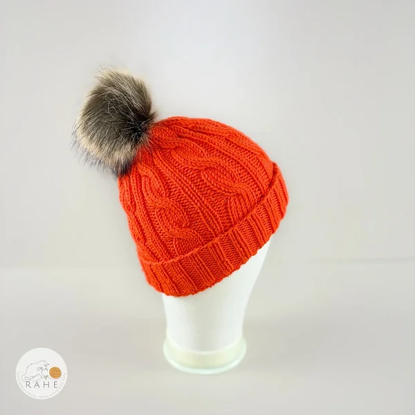 Right side view of an orange hand-knitted merino wool children’s hat with faux fur pom-pom by RAHEcraft