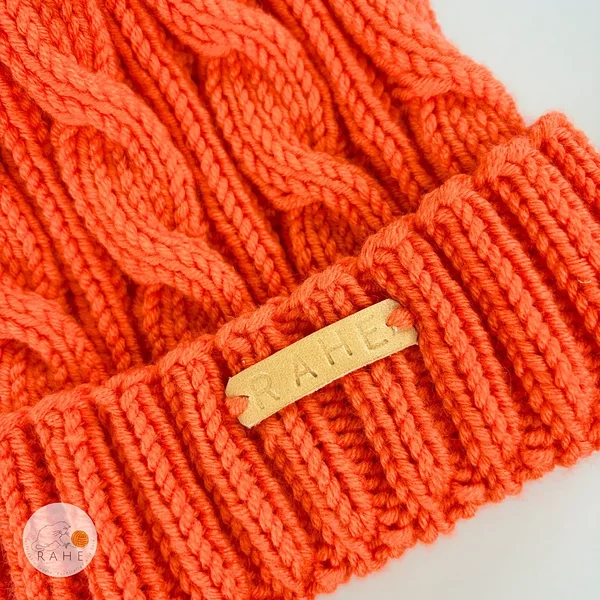 Close-up of the cable knit texture and ribbed brim on an orange hand-knitted merino wool children’s hat by RAHEcraft