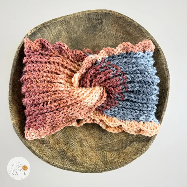 Hand-knitted wide acrylic headband laid flat on a wooden plate, soft blue and peach gradient with twisted front design, ribbed knit texture and hand-crocheted scalloped edge, RAHEcraft